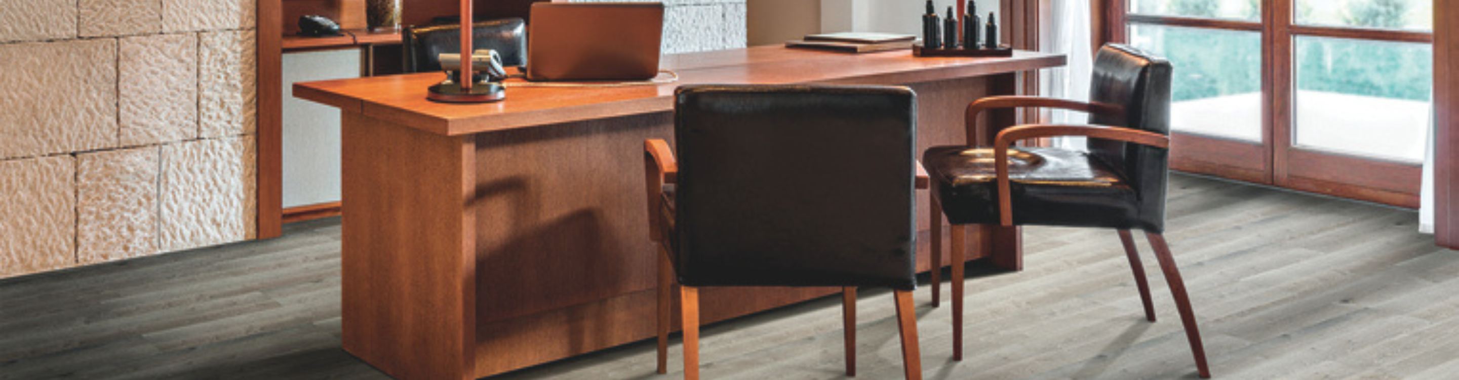 Office space with gray wood flooring, a large wooden desk, black leather chairs, textured stone walls, and warm lighting, showcasing a sophisticated and professional interior design.